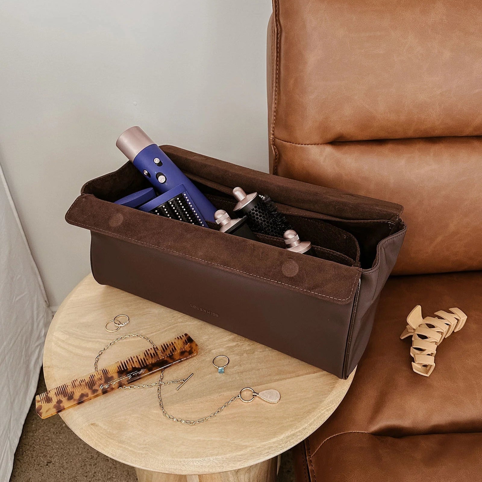 Brown leather bag with hair styling tools on a wooden stool next to a brown leather chair.